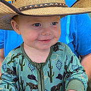 Tyrus is registered to the contest to win money with this photo: toddler, child, cowboy_hat, smile, blue_eyes, green_onesie, horse_pattern, cactus_pattern, zipper, long_sleeves, person, face, hand, background_person, blue_shirt, indoor, portrait, cute, happy, baby