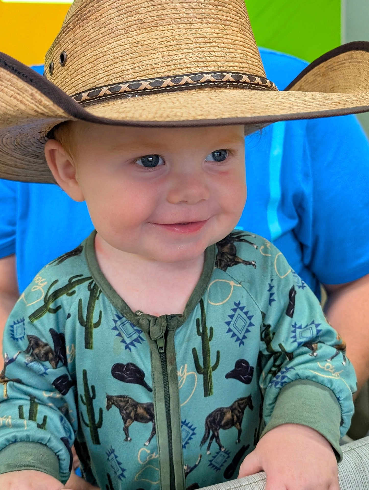Tyrus is registered to the contest to win money with this photo: toddler, child, cowboy_hat, smile, blue_eyes, green_onesie, horse_pattern, cactus_pattern, zipper, long_sleeves, person, face, hand, background_person, blue_shirt, indoor, portrait, cute, happy, baby