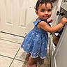 toddler, child, blue_dress, barefoot, bathroom, cabinet, curious, smile, face, person, floor, tile, door, rug, earrings, hairclip, standing, indoor, young_child, cabinet_handle