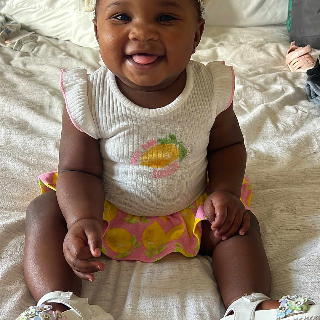 Zanai is registered to the contest to win money with this photo: baby, bed, bows, child, curly_hair, cute, footwear, happy, indoor, infant, lemon_print, person, pink, playful, sitting, skirt, smiling, tongue_out, white_shoes, white_top