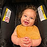 baby, child, car_seat, smile, curly_hair, blue_eyes, yellow_bib, happy, infant, seatbelt, clothing, hands, portrait, cute, indoor, person, toddler, sitting, closeup, face