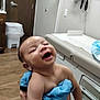 Cobee is registered to the contest to win money with this photo: baby, infant, smiling, laughing, blue_cloth, medical_room, doctor_office, wood_floor, trash_bin, medical_equipment, door, cabinet, person, happy, cute, child, skin, holding, interior, healthcare
