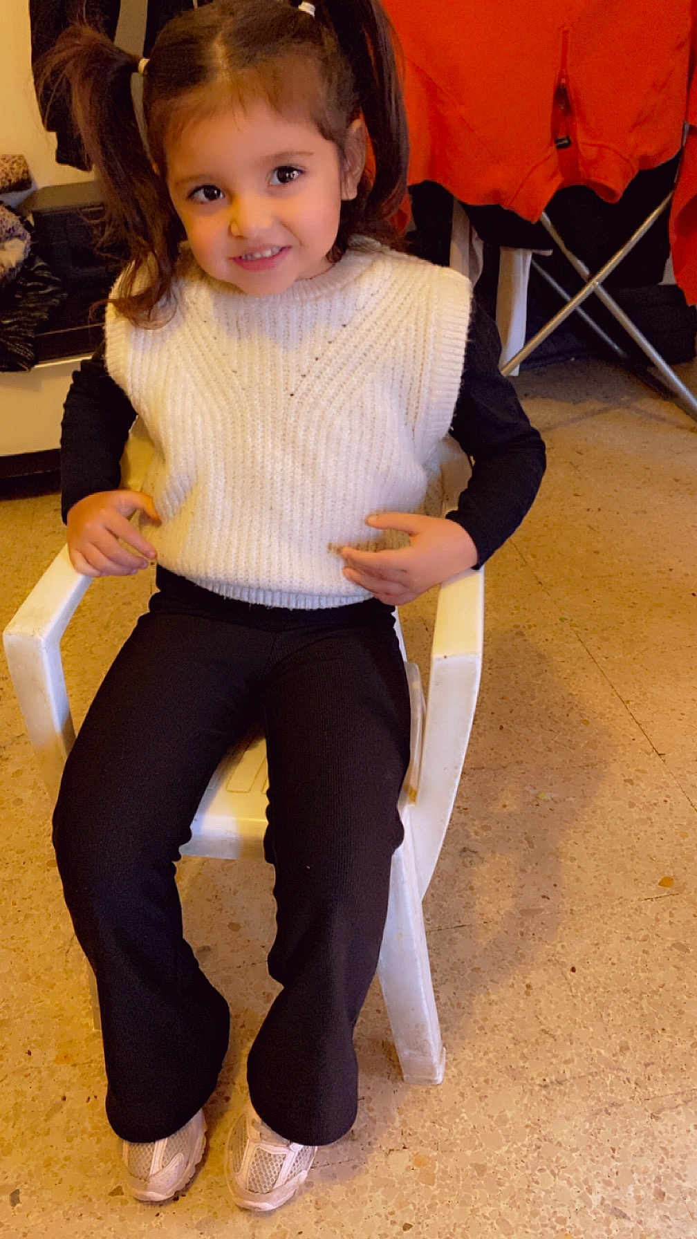 Maelya a rejoint le concours — aidez-le/la à gagner de superbes lots ! child, girl, smiling, pigtails, white_vest, black_shirt, black_pants, sneakers, plastic_chair, indoor, floor, clothing, person, happy, seated, portrait, casual, young_child, home, warm_lighting