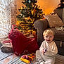 toddler, child, toy, christmas_tree, holiday, presents, indoor, carpet, sofa, cushion, curtain, plush_toy, warm_lighting, decorations, smiling, blonde_hair, blue_eyes, festive, winter, cozy
