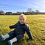 toddler, child, grass, field, outdoor, sunlight, blue_sky, jacket, rain_boots, smiling, blonde_hair, trees, houses, daylight, casual_clothing, sitting, nature, happy, park, playful