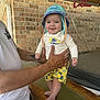 Emmett is registered to the contest to win money with this photo: baby, child, smiling, hat, tropical_clothing, yellow_shorts, adult_hands, brick_wall, wooden_surface, net, indoor, person, cute, happy, foot, standing, casual_clothing, summer, headwear, portrait