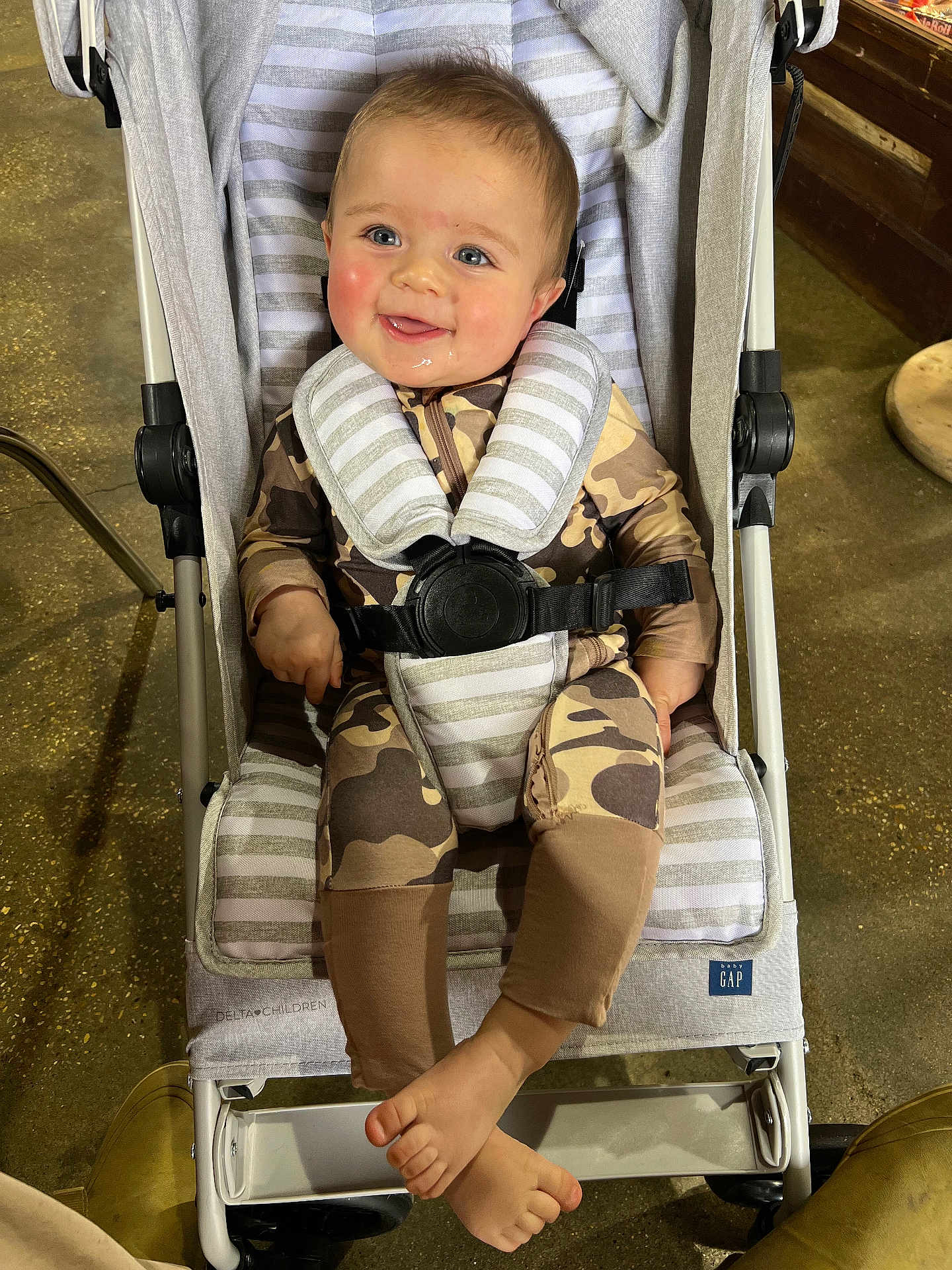 Emmett is registered to the contest to win money with this photo: baby, stroller, child, camo_onesie, harness, smiling, drool, blue_eyes, rosy_cheeks, indoor, floor, footwear, striped_padding, happy, seated, infant, baby_gap, clothing, legs, toes